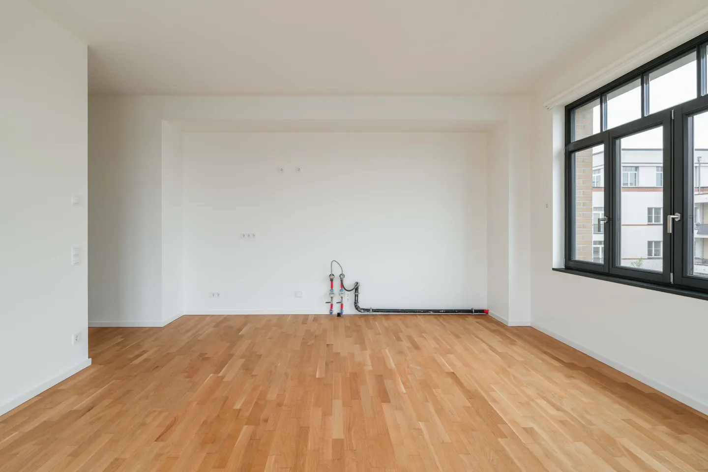 Bright, empty room with light wood floors, white walls, and a black-framed window showing an apartment building outside.