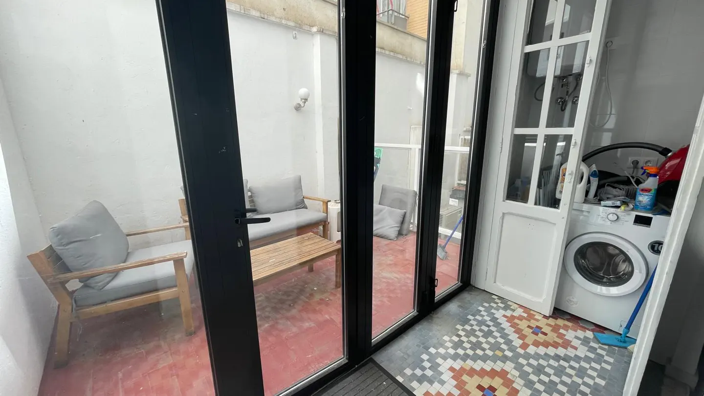 A sunroom with black framed glass doors, a seating area with gray cushions, and a laundry area with a white washing machine.