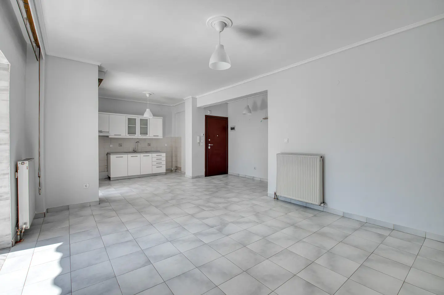 Bright, empty apartment with white walls, tile floors, and a small kitchen. A dark wood door is visible in the background.