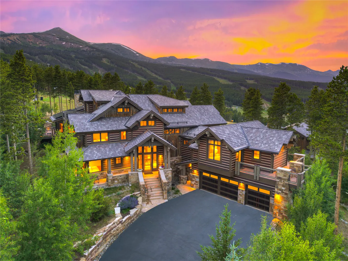 A large log cabin home with a gray roof sits in front of a mountain range at sunset.