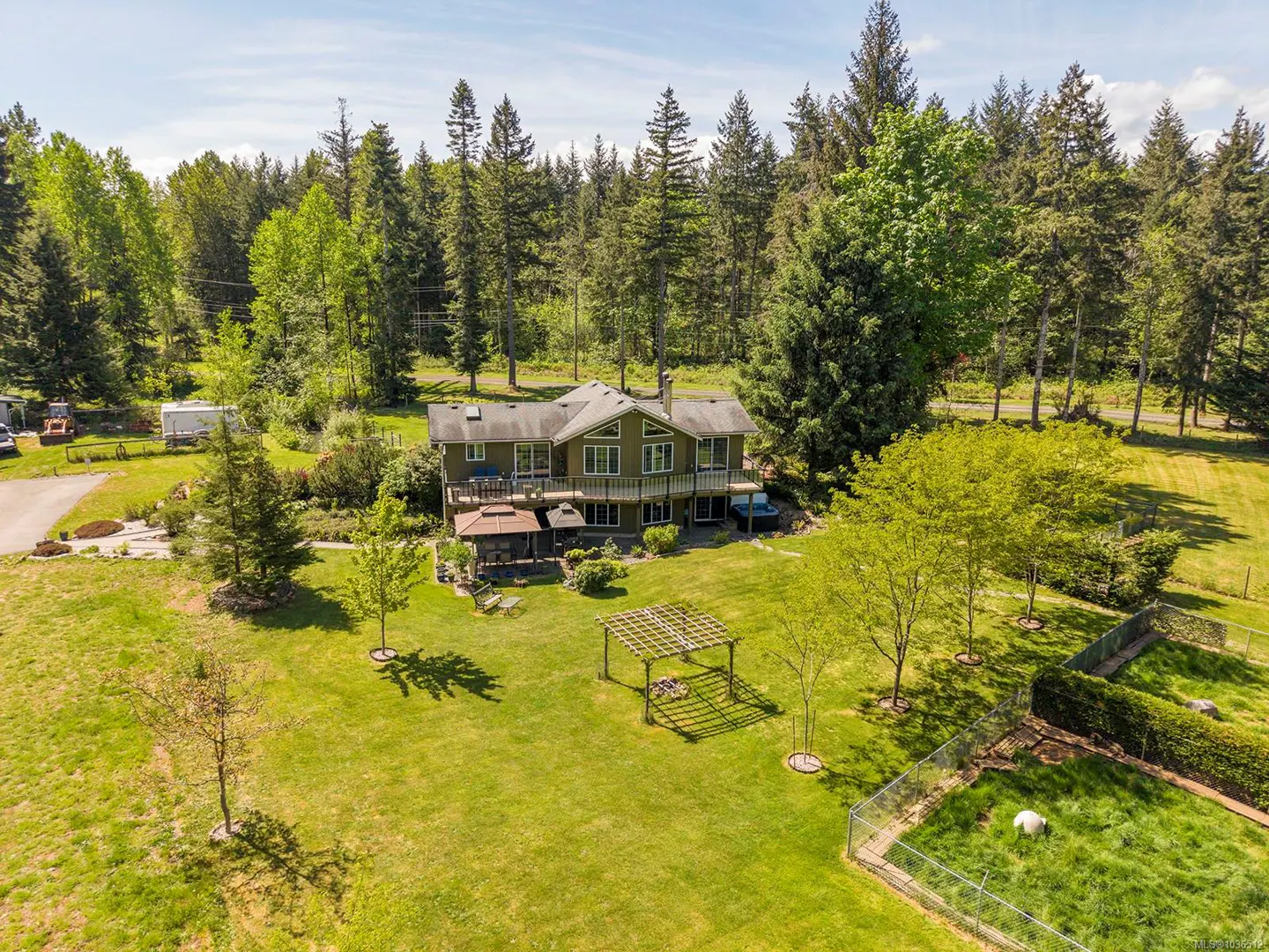 Aerial view of a two-story olive green house with a deck, surrounded by a lush green lawn, trees, and a forest backdrop.
