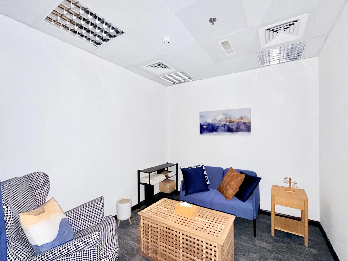 Small office lounge with a blue sofa, houndstooth armchair, wooden coffee table, and abstract wall art on white walls.