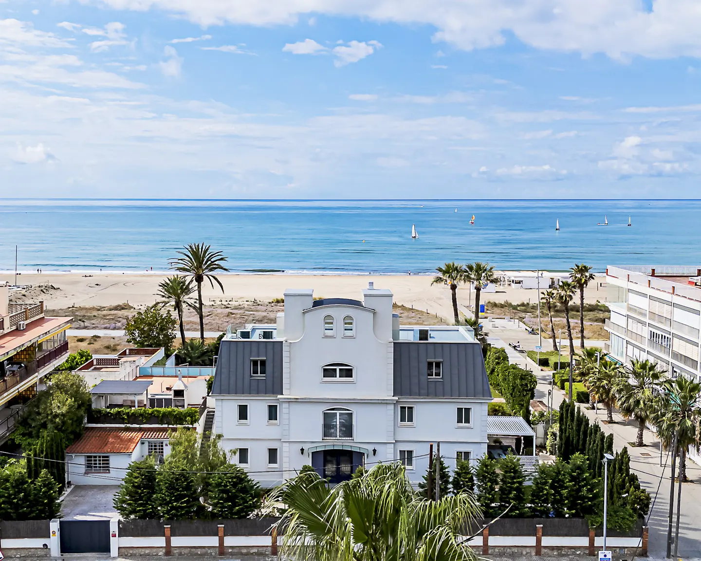 View of a white, multi-story house with a gray roof, palm trees, and the ocean in the background.