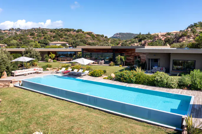 Aerial view of a modern home with a long, rectangular pool, lounge chairs, and green lawn against a backdrop of trees and a blue sky.