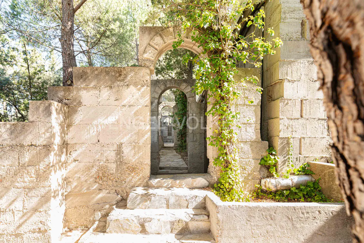 Stone arches lead to a path in a garden. Green vines grow on the stone. Steps lead up to the first archway.