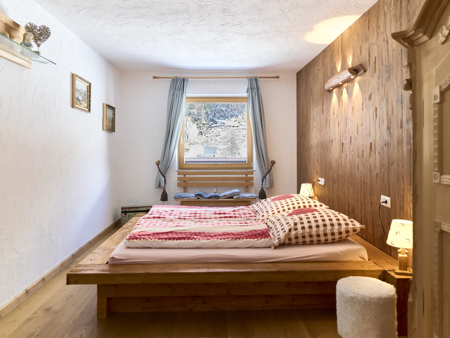 Bedroom with a wooden platform bed, red and white bedding, and a window with blue curtains. A wooden wall with a unique light fixture adds rustic charm.