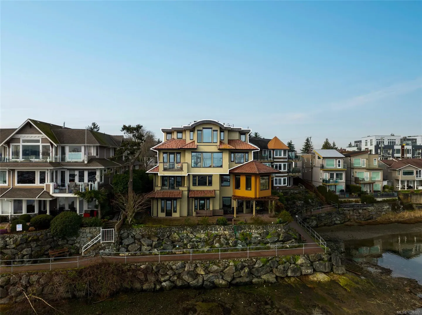 A three-story beige house with a red tile roof sits on a rocky shoreline under a blue sky.