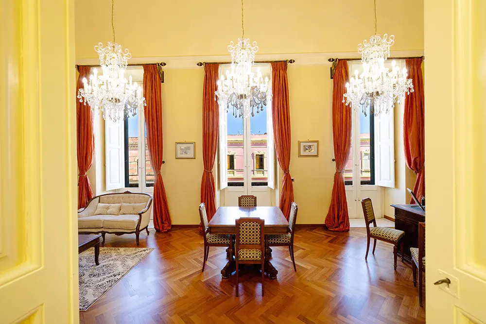 A bright room with yellow walls, three chandeliers, and a wooden floor. There is a table with chairs, a sofa, and red curtains.