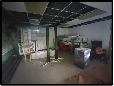 A cluttered room with exposed ceiling tiles, furniture, mattresses, and office chairs. The walls are white, and the floor is tiled.