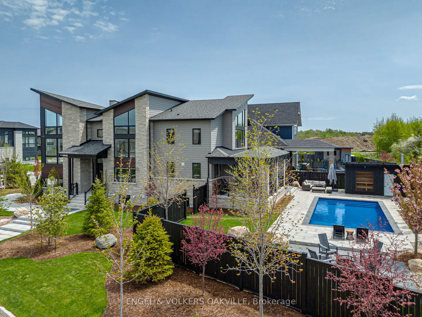 Modern two-story house with a pool. The house is gray with stone accents and large windows. The pool is surrounded by a patio and lounge furniture.