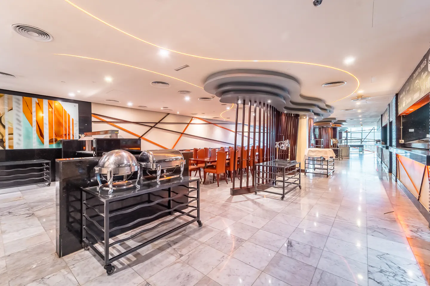 A bright, modern restaurant interior with buffet tables, marble floors, and orange accents.