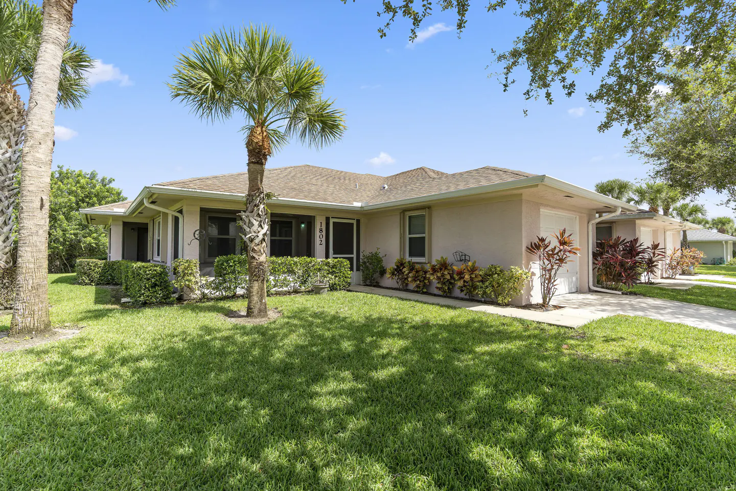 Single-story light pink house with a palm tree in the front yard, green lawn, and a white garage door under a blue sky.