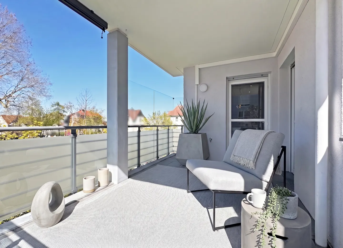 A modern balcony with a gray chair, blanket, and side table with a plant. A large potted plant sits near the glass railing. Outside is a blue sky.