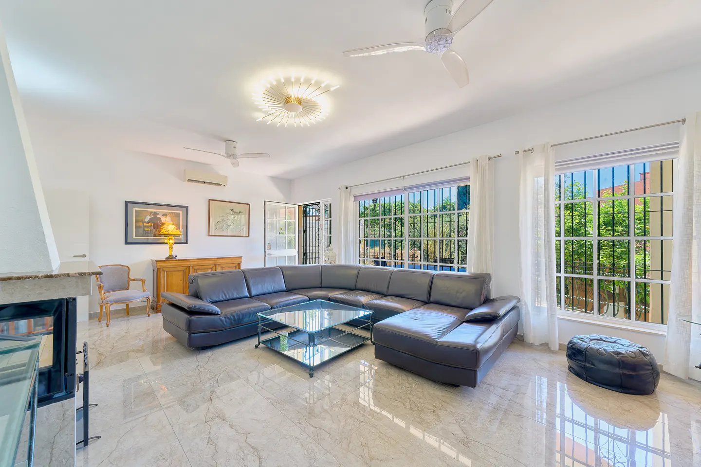 Bright living room with a large black leather sectional sofa, glass coffee table, and marble floors. Windows overlook lush greenery.