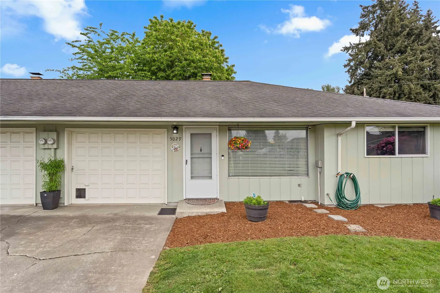Exterior of a one-story, light green home with a white garage door and front door. A hanging flower basket is near the front door.
