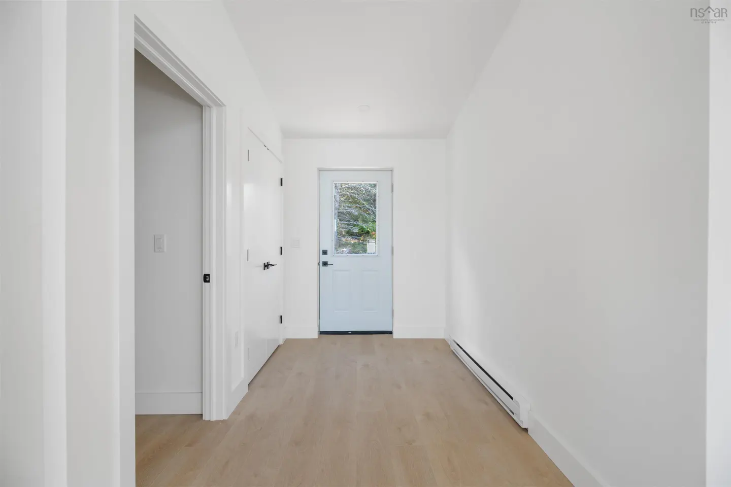 Bright hallway with light wood floors, white walls, and a white door with a window to the outside.