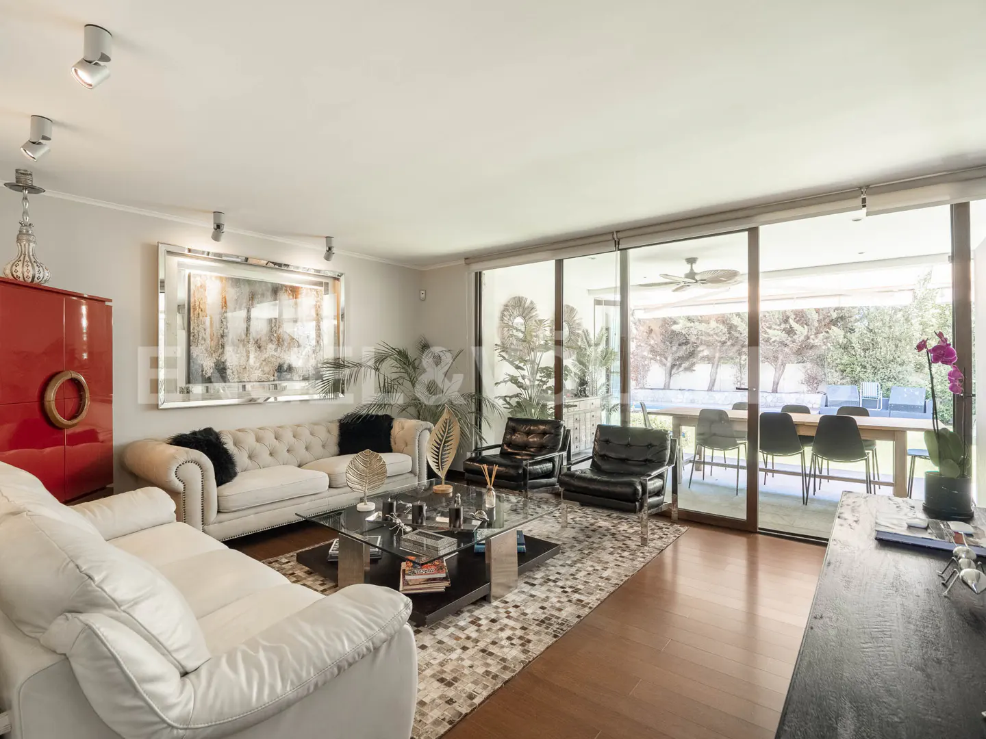 Bright living room with white sofas, black chairs, and a glass coffee table. Large windows overlook an outdoor dining area.