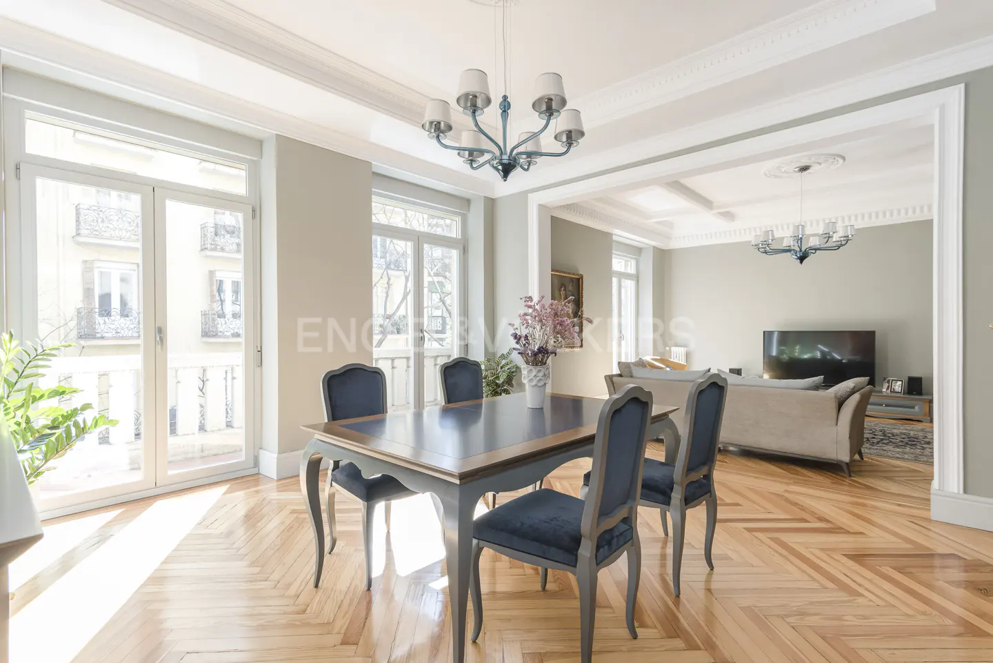 A dining room with a dark wood table, blue chairs, and a chandelier. A living room with a sofa and TV is visible in the background.