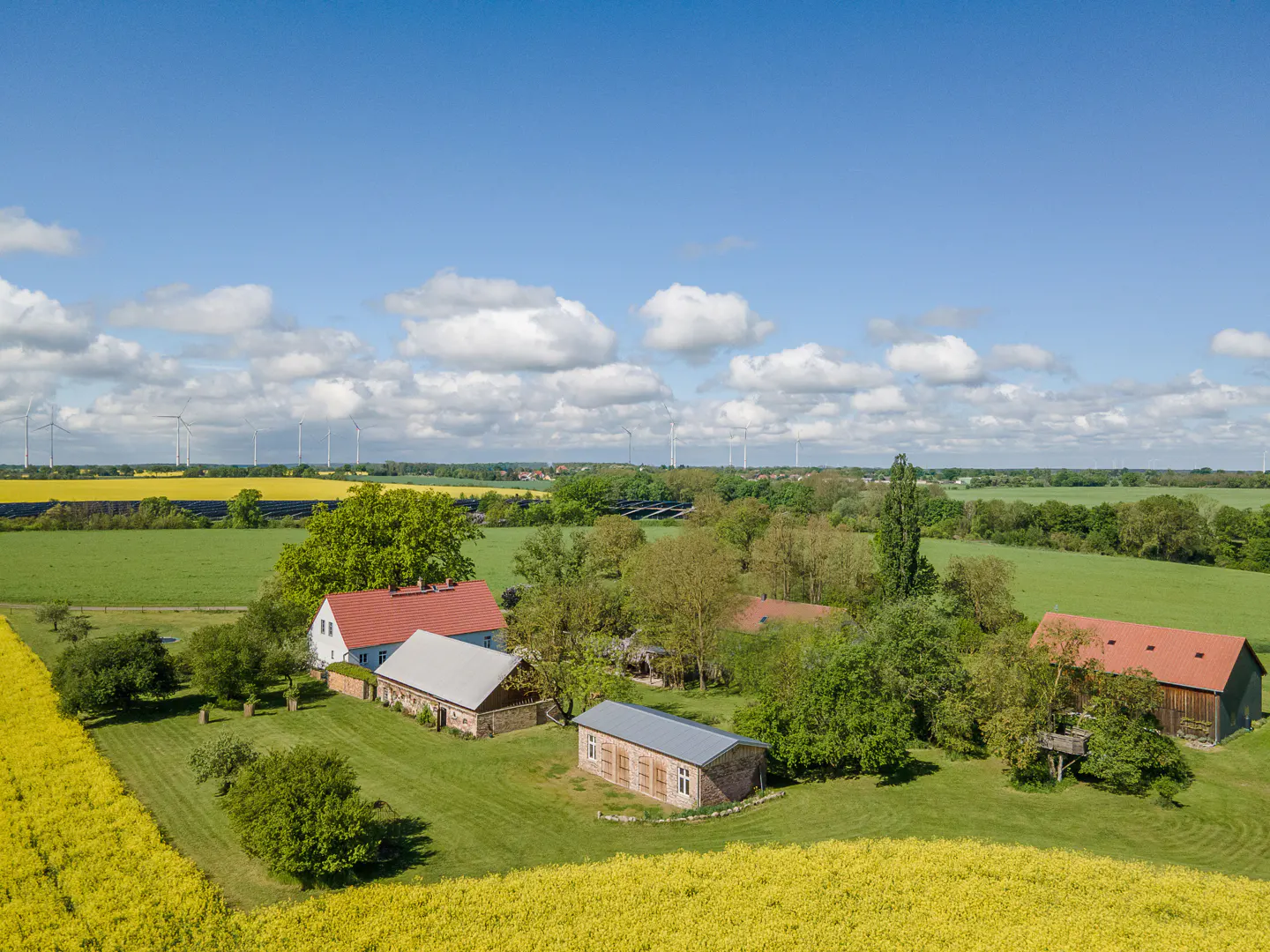 Aerial view of a rural property with several buildings, green lawns, trees, and yellow fields under a blue sky with white clouds. Wind turbines are visible in the distance.