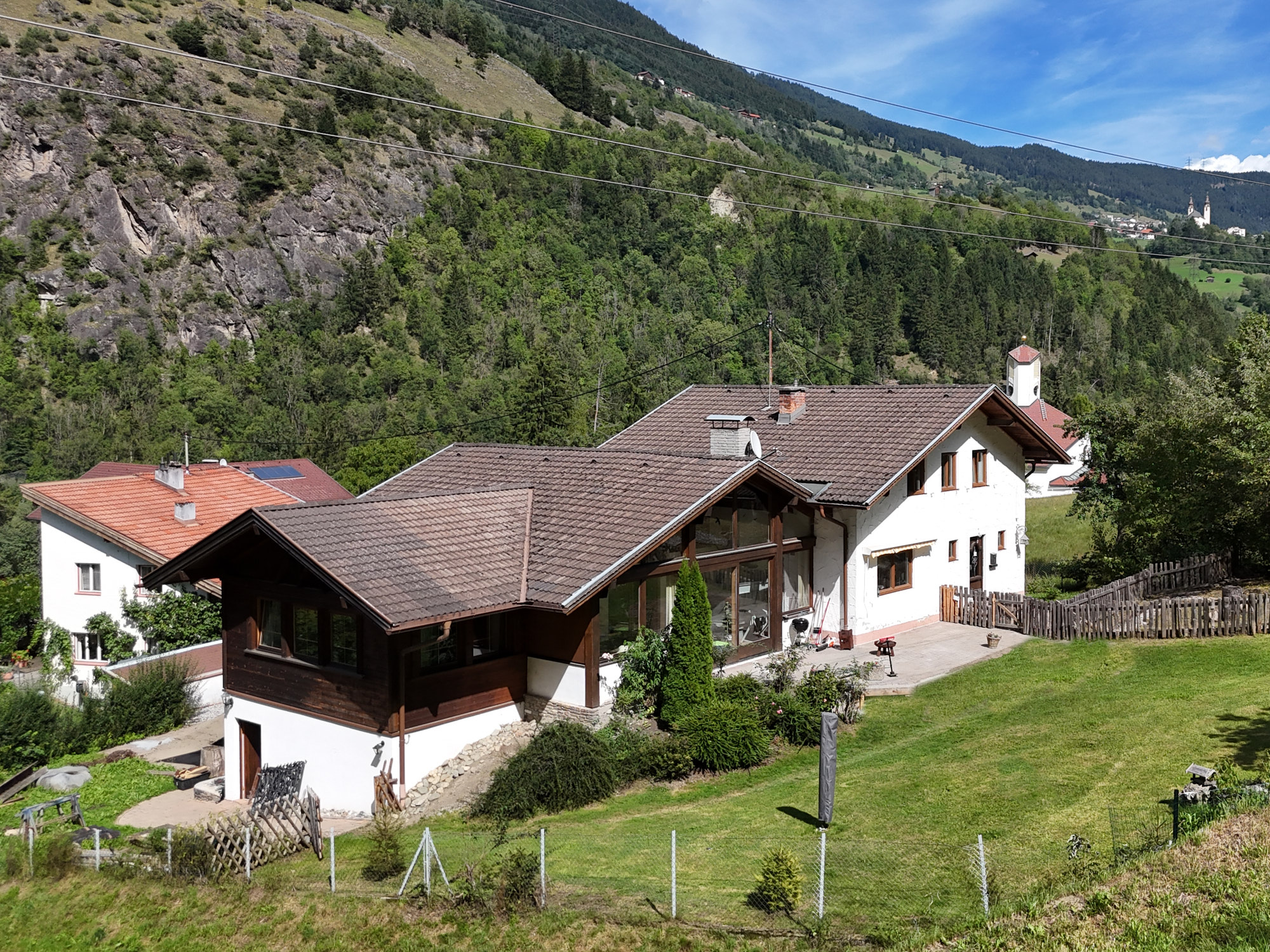 A white house with a brown roof and large windows sits on a green lawn against a backdrop of forested mountains. [AI-generated text]