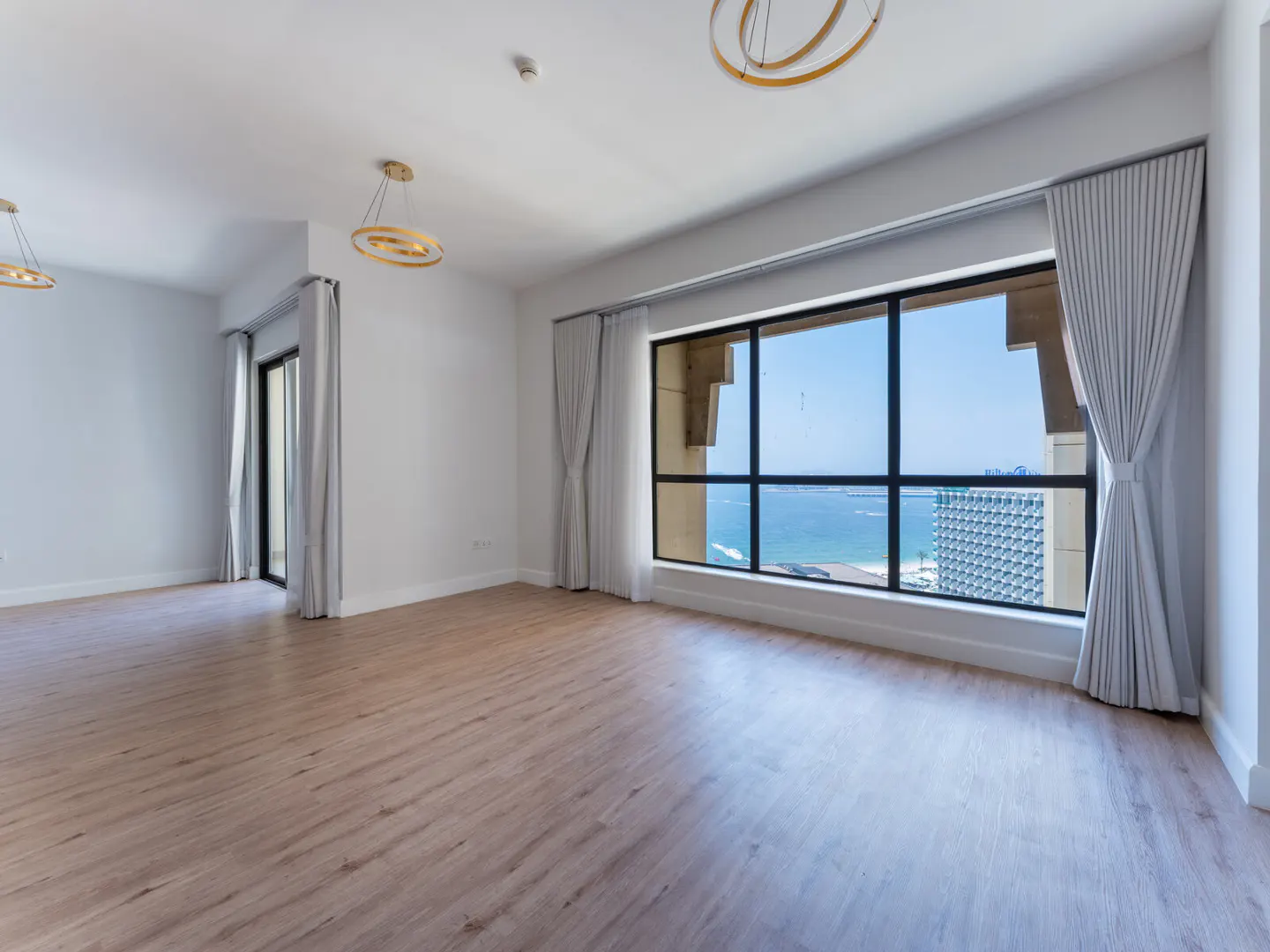 An empty room with light wood floors, white walls, and a large window with ocean views. Gray curtains frame the window. Two modern chandeliers hang from the ceiling.