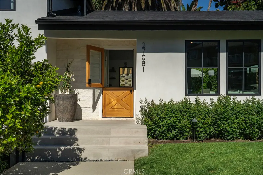 Exterior of a white house with a wooden Dutch door, black framed windows, and green landscaping.