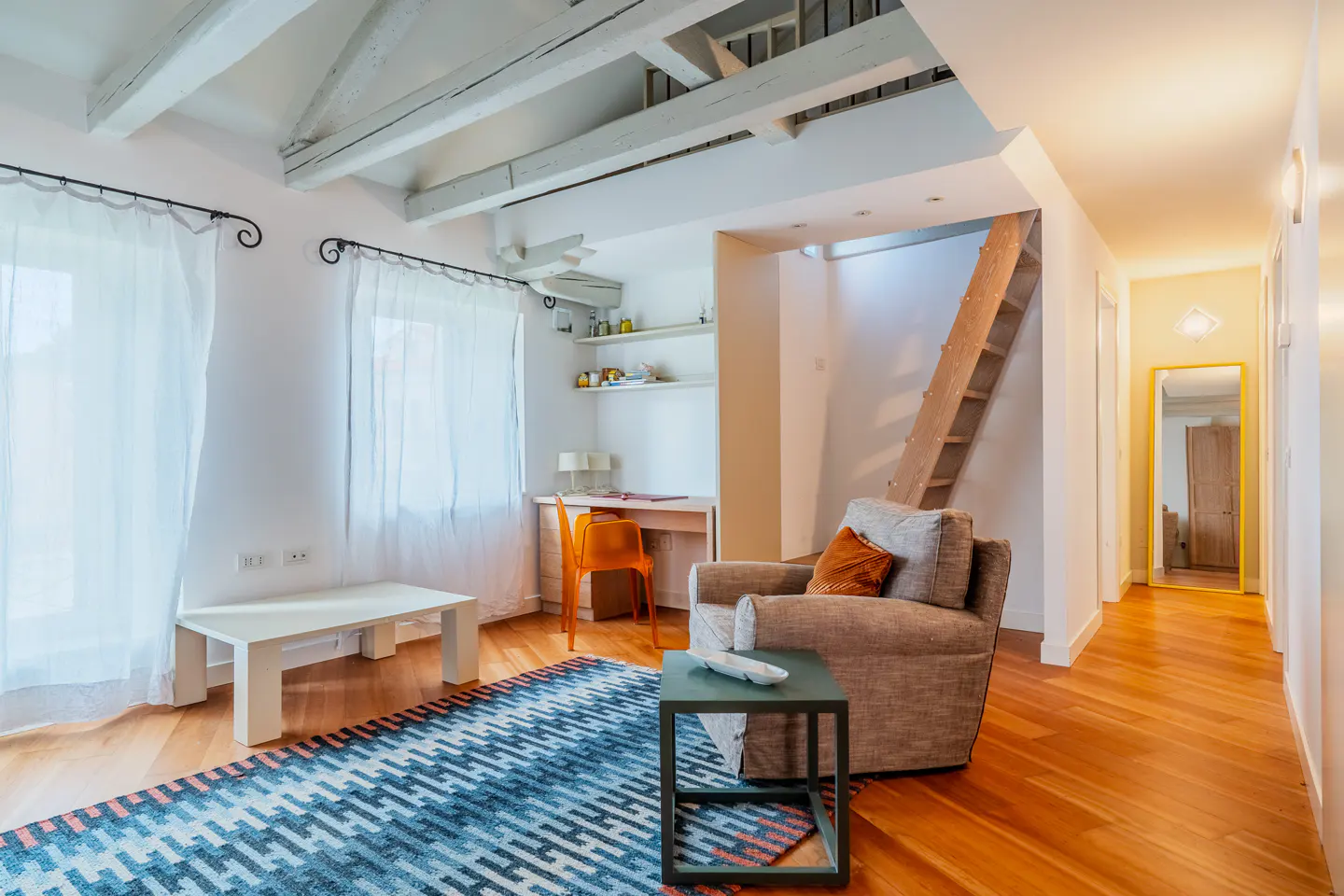 Bright living room with wood floors, blue patterned rug, grey armchair, orange desk chair, and wooden stairs to a loft.