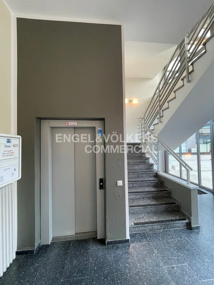 Interior view of a commercial building lobby with a gray elevator, stairs with metal railings, and a black speckled floor.