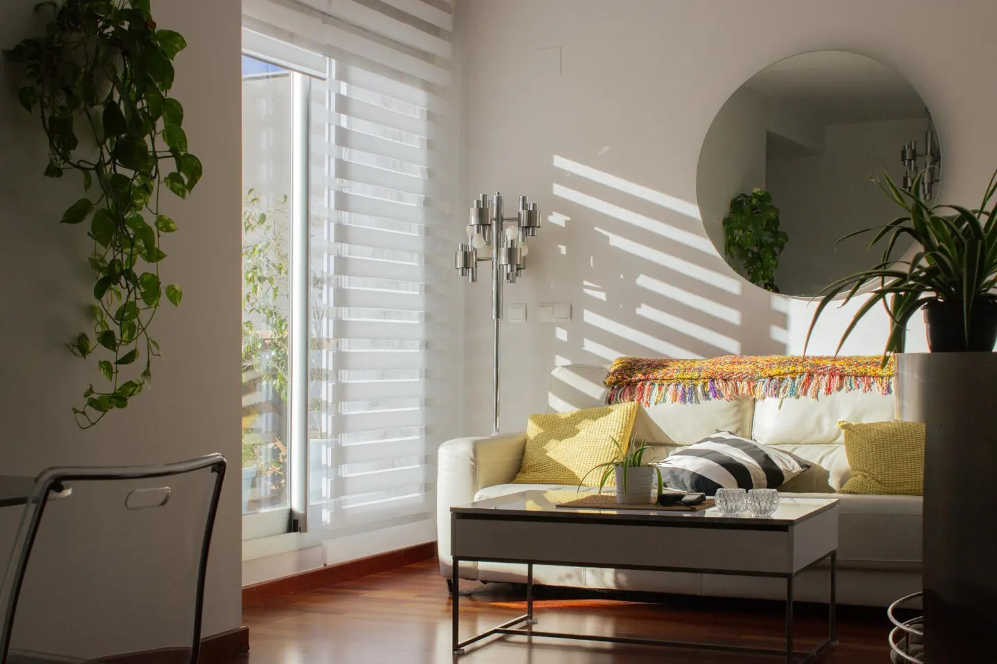 Bright living room with white sofa, yellow pillows, and striped cushion. Sunlight streams through window blinds. Round mirror on the wall.