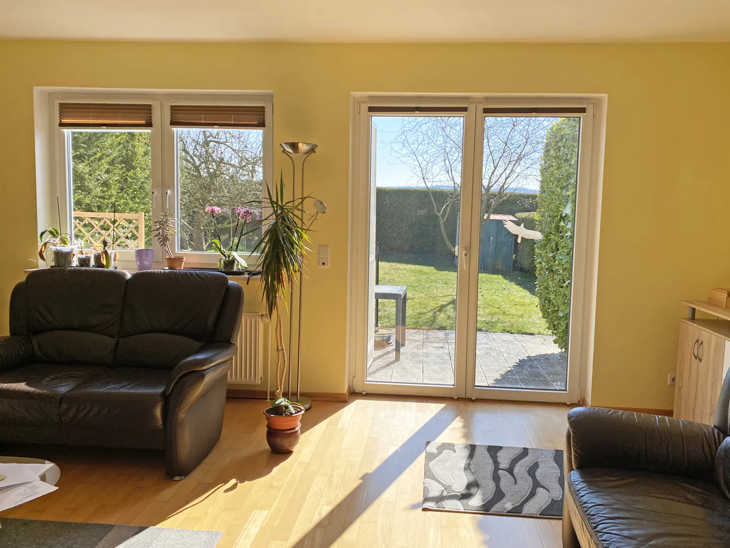 Sunny living room with yellow walls, dark leather sofas, and a sliding glass door to a green backyard. Windows with plants and blinds.