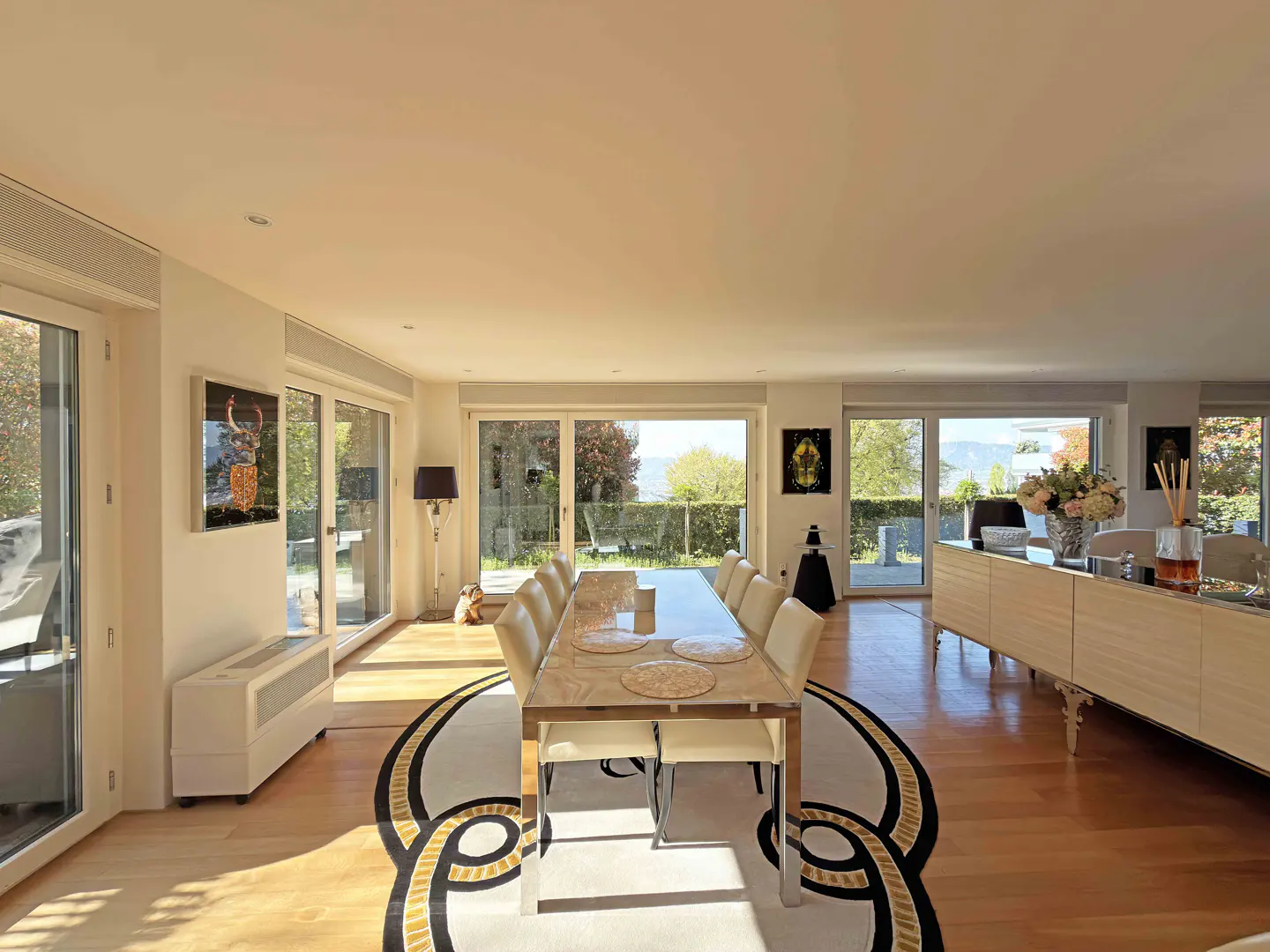 Bright, modern dining room with a long table, cream chairs, and a decorative rug. Large windows offer a view of greenery.