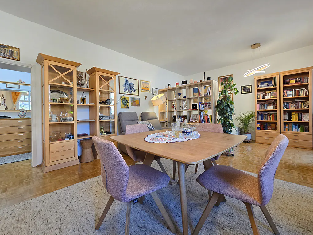 A bright living room with a wooden table, four pink chairs, and a white rug. Bookshelves and artwork decorate the walls.