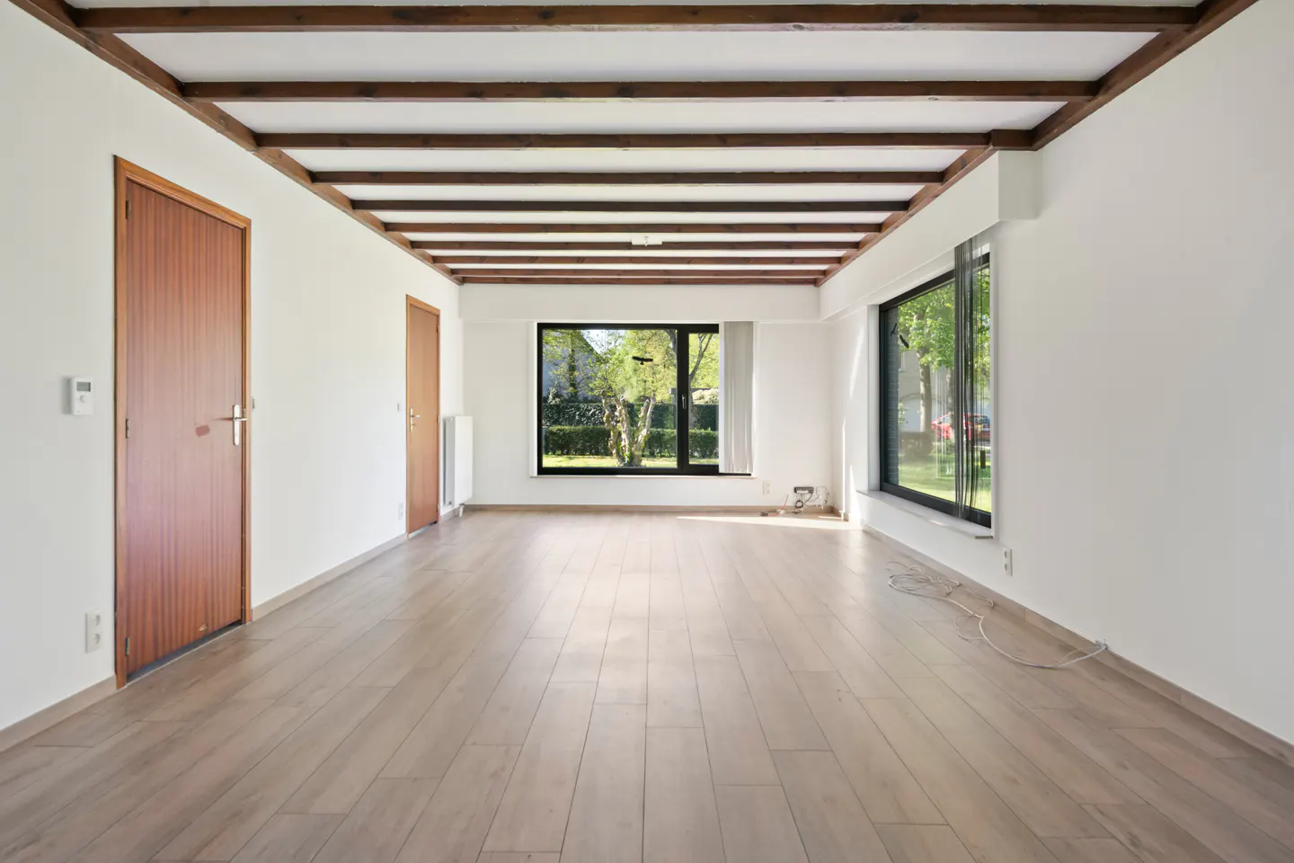 Bright, empty room with wood floors, white walls, and exposed wood beams on the ceiling. Large windows offer a view of a green yard.