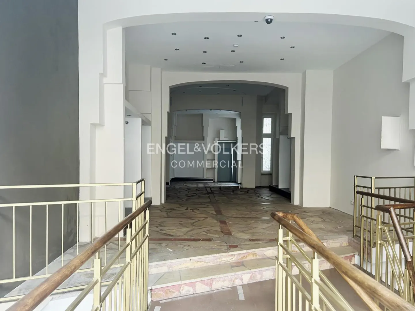 Interior view of a commercial space with stone floors, white walls, and metal railings. Engel & Völkers Commercial logo is visible.