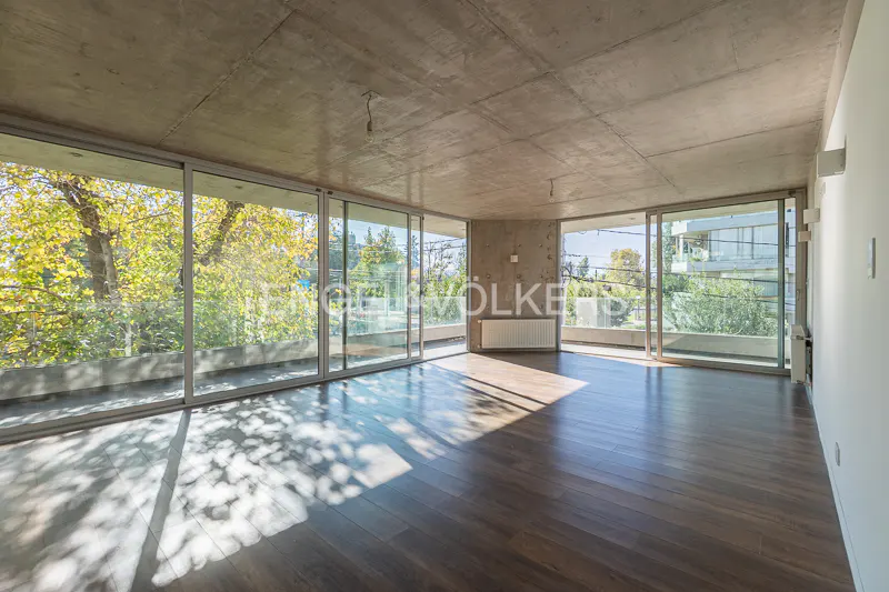 Empty room with dark wood floors, concrete ceiling, and large windows showing trees outside.