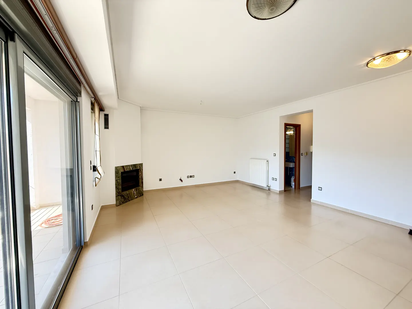 Bright, empty room with white walls, beige tile floor, and a green marble fireplace. Sliding glass doors lead to a balcony.
