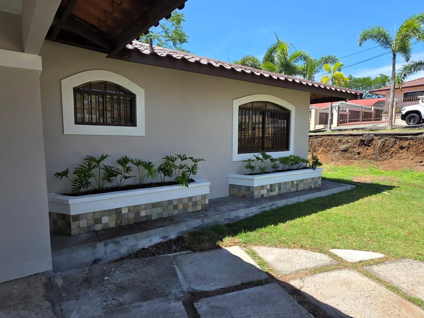 Exterior view of a beige house with arched windows, brown trim, and planter boxes filled with green plants. A lawn and palm trees are visible.