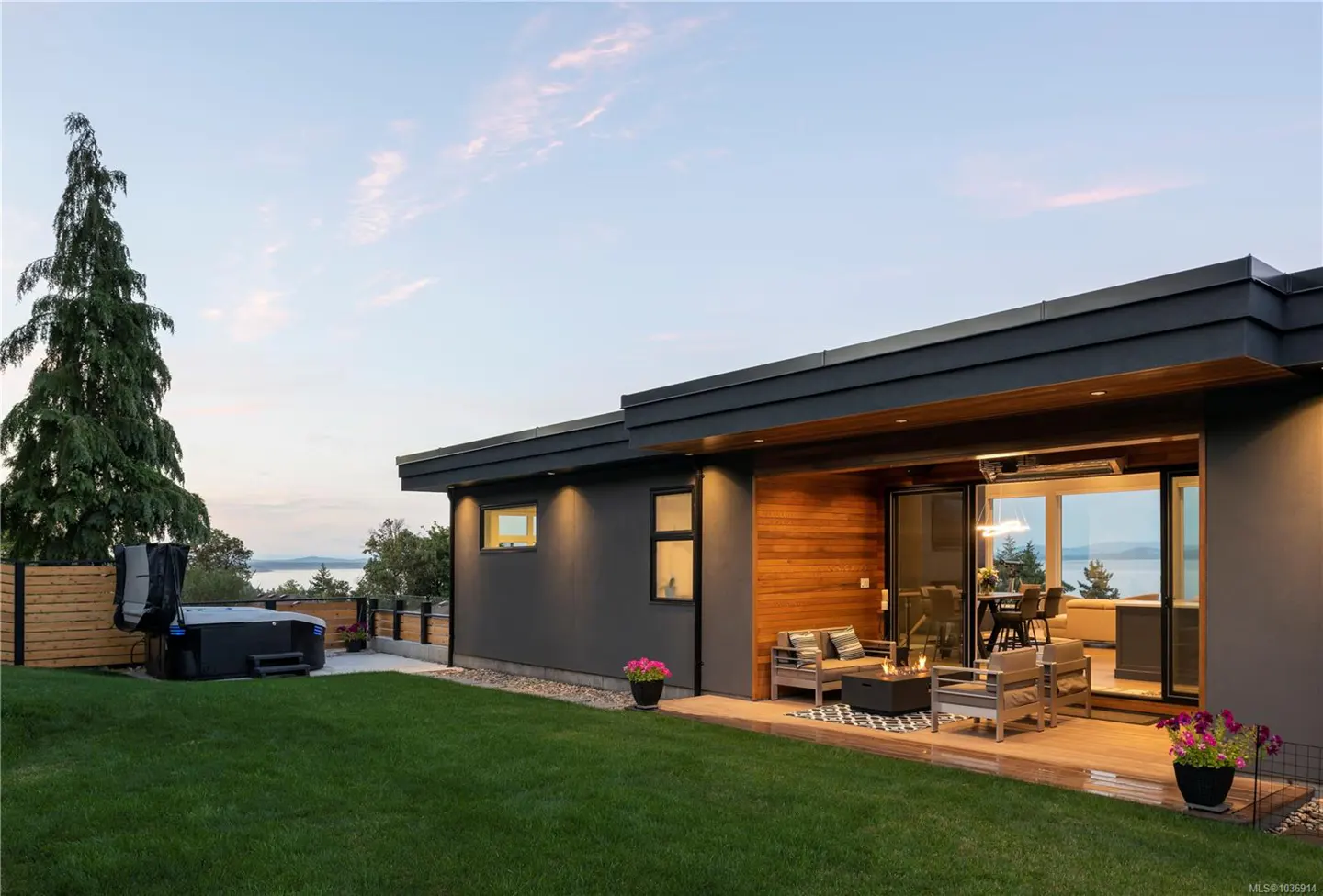 Modern home exterior with a gray facade, wood accents, and a patio with seating overlooking a green lawn and distant water view.