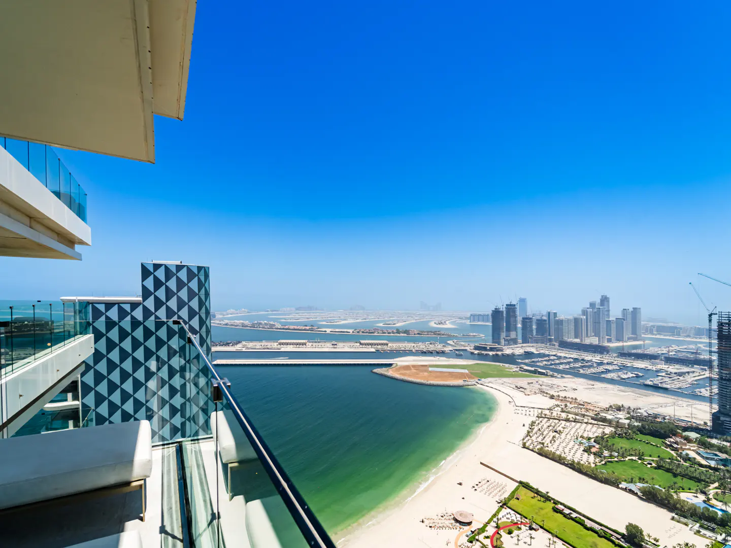 High-rise balcony with white lounge chairs overlooking a turquoise beach, marina, and distant city skyline under blue sky.
