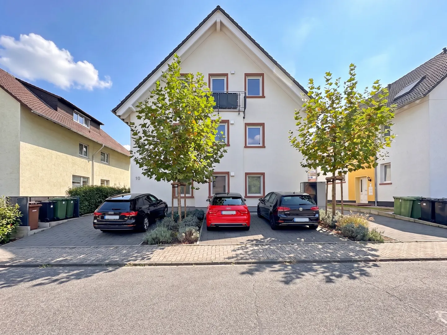 A white two-story house with three cars parked in front, two black and one red, under a blue sky.