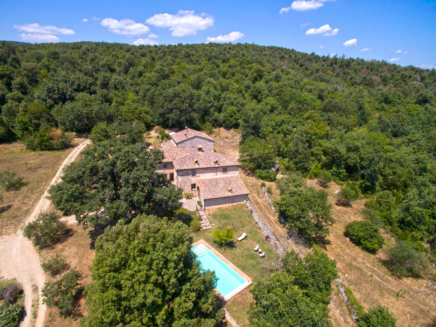 Aerial view of a stone house with a red tile roof, a blue pool, and a lush green forest backdrop.