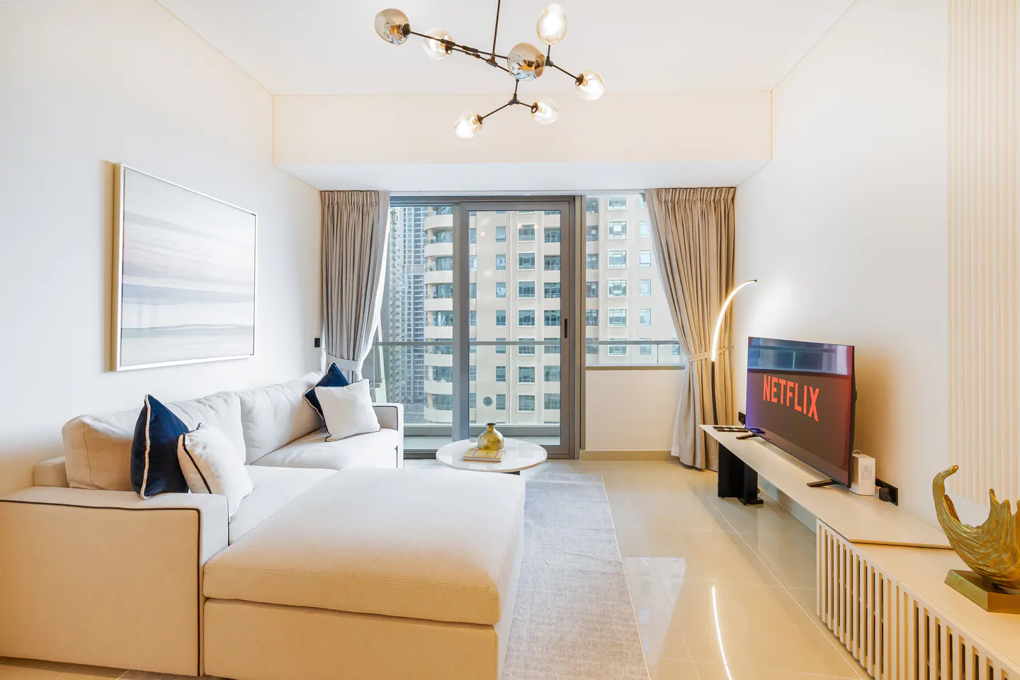 Bright living room with a white sectional sofa, a TV showing Netflix, and a city view through a sliding glass door.