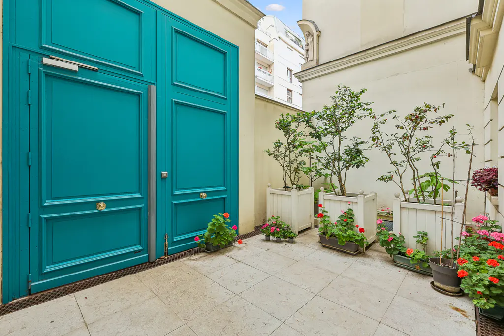A tiled patio features a large, bright teal double door and several white planters filled with green shrubs and flowers.