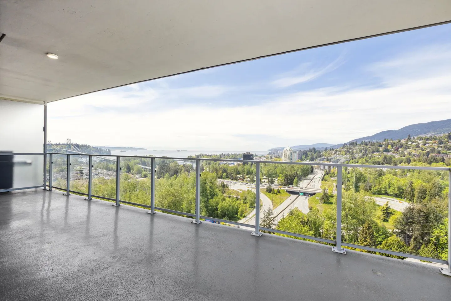 View from a condo balcony with a glass railing overlooking a city, trees, and mountains. Blue sky with white clouds.