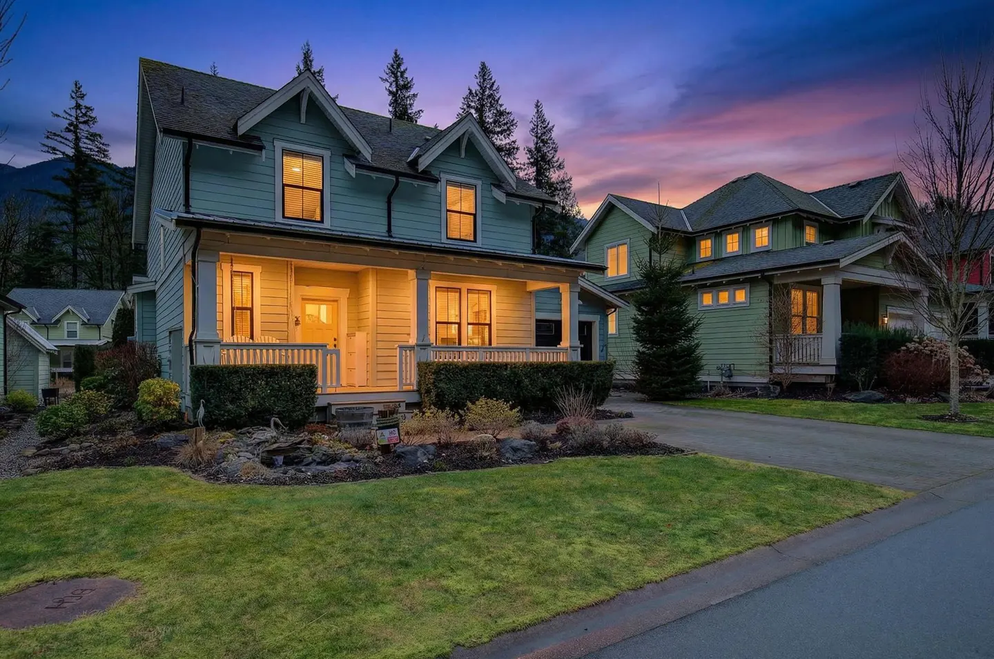 Light blue two-story craftsman home with a glowing front porch at dusk, set against a vibrant purple and orange sunset.