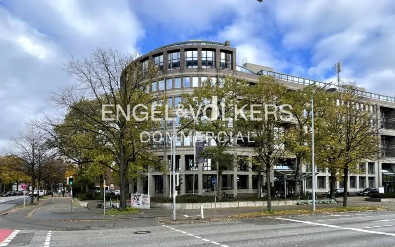 Exterior of a modern, multi-story commercial building with "Engel & Völkers Commercial" signage, surrounded by trees and a city street.