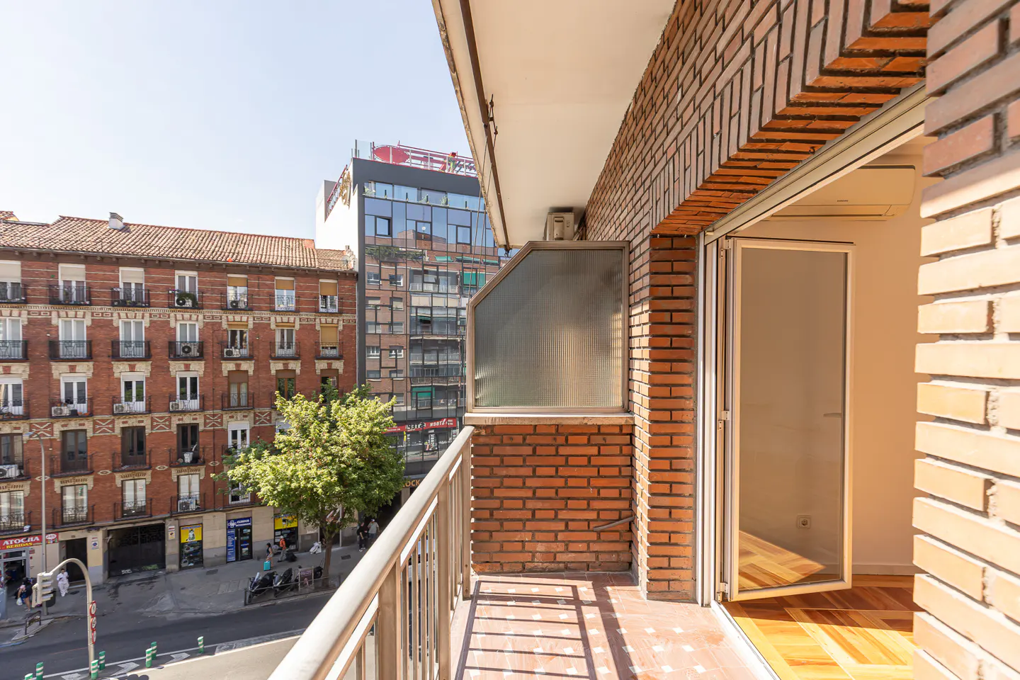 View from a brick balcony with metal railing overlooking a city street with buildings and pedestrians.