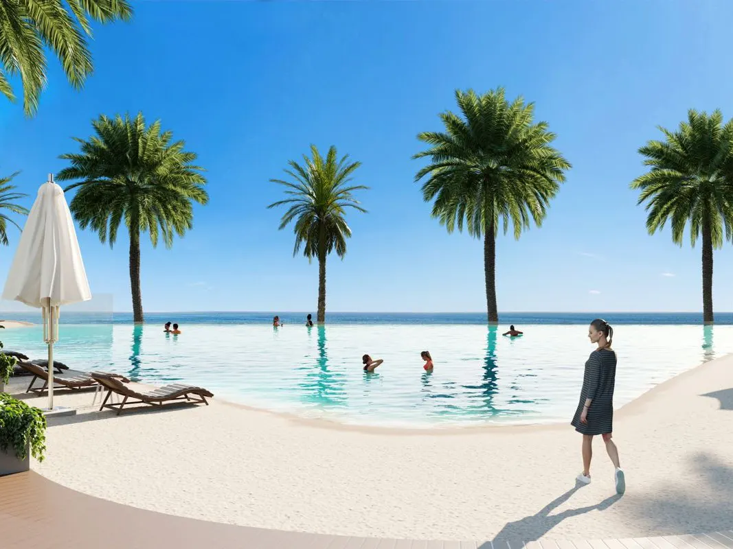 Infinity pool with palm trees, blue sky, and ocean view. Woman in dress walks on the sand.