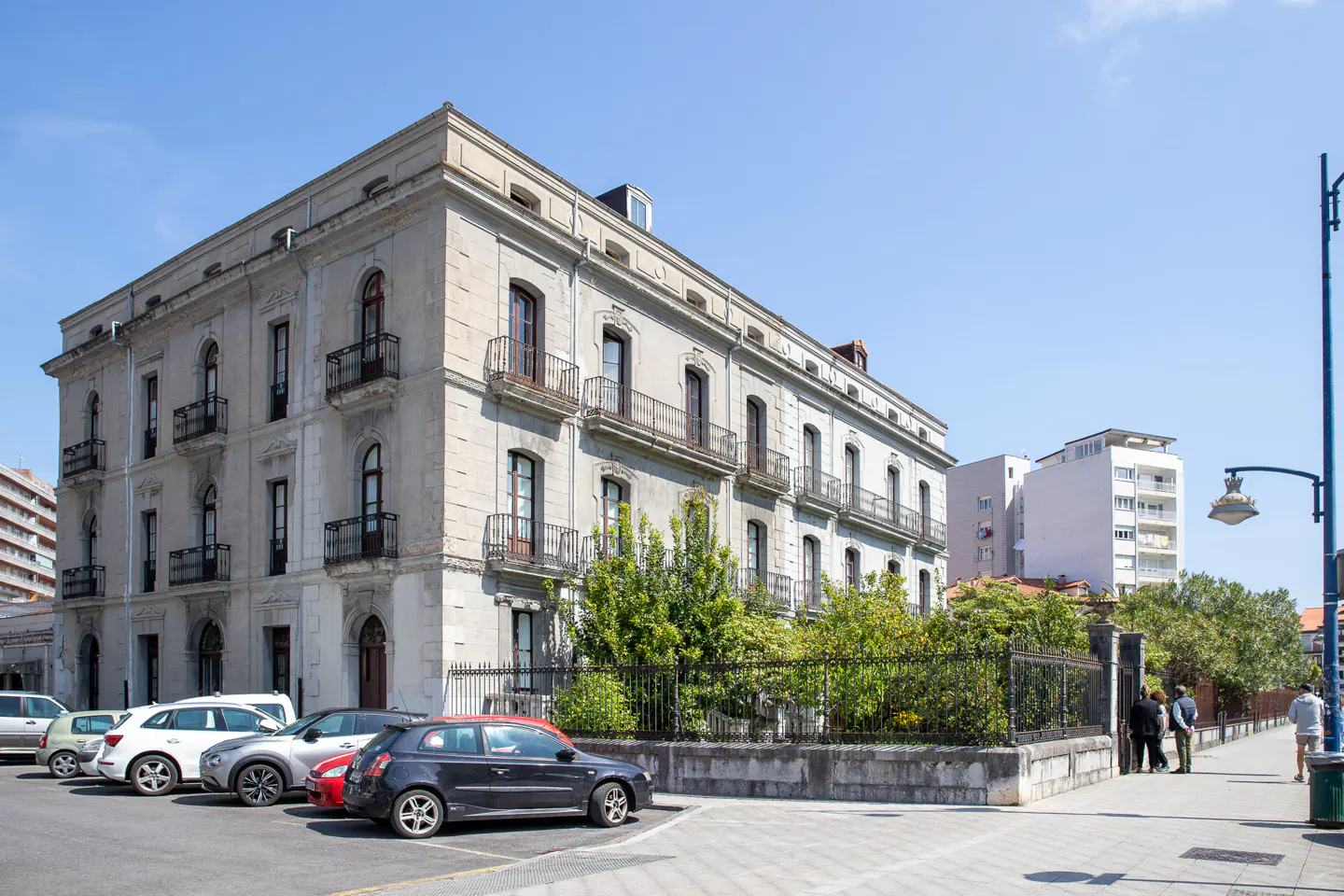 A large, light gray building with black balconies and arched windows, cars parked in front, and people on the sidewalk.