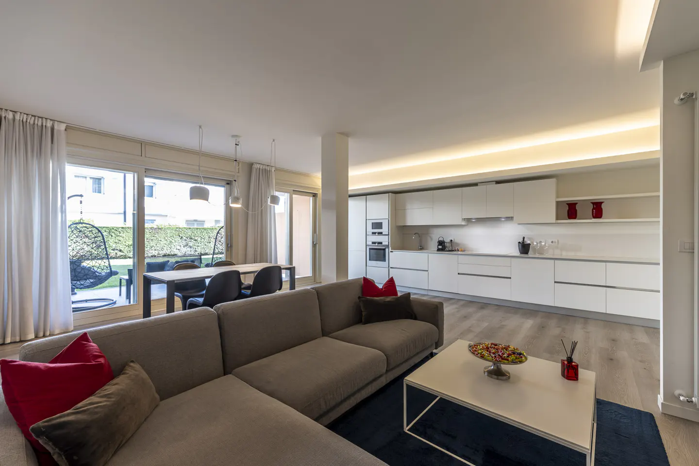 Open-concept living space with a gray sectional sofa, white kitchen cabinets, and a dining table near a window with a garden view.
